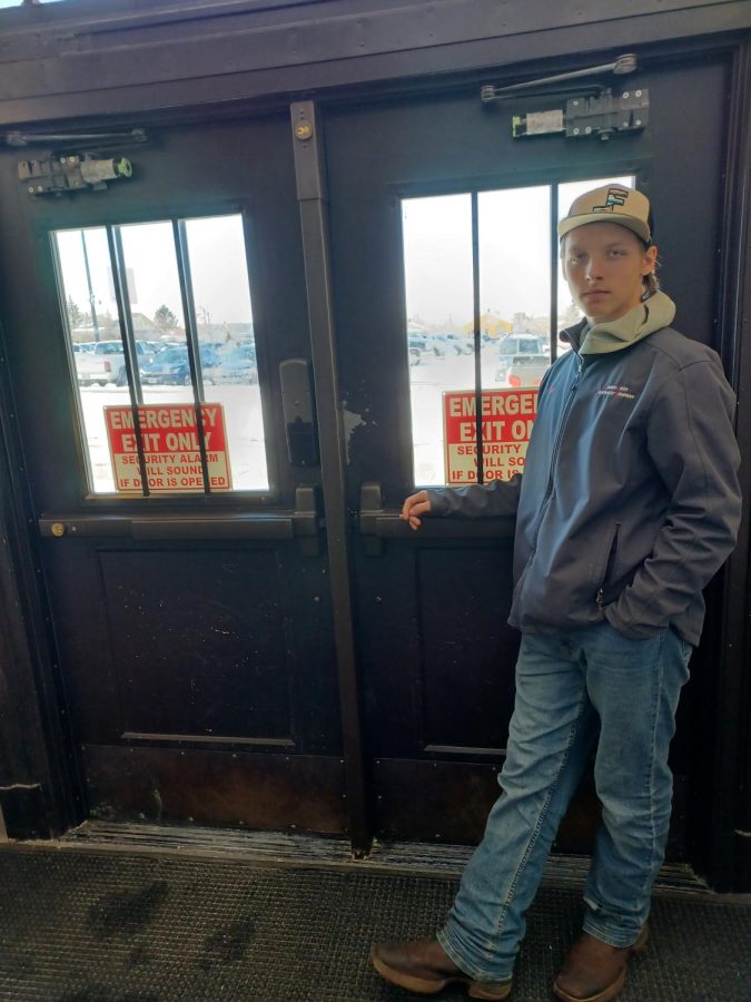 An NC student stands in front of doors with emergency exit signs. Student is wearing jeans, hat, and blue coat. Cars visible outside of windows in student parking lot.