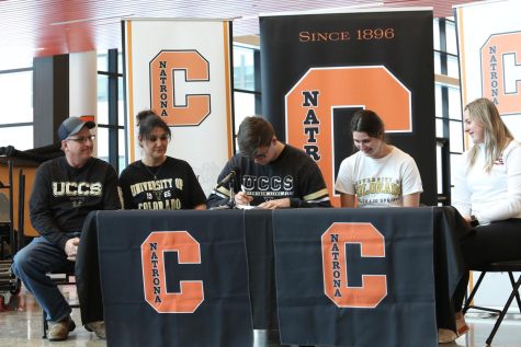 Santistevan, wearing UCCS sweatshirt, with NCHS orange and black logo banner in background and family surrounding him, looks down to sign his letter of intent.