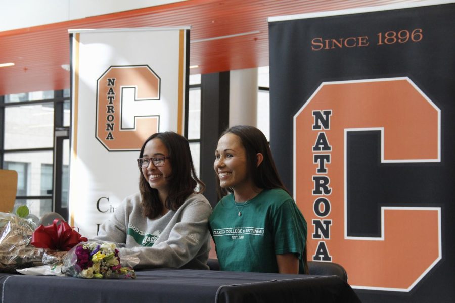 Baker and her mother sit at a table after signing. Orange and black school logo posters in background.