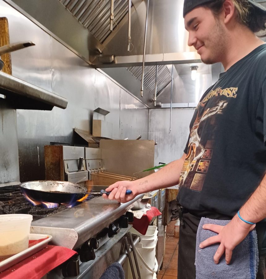 A high school student in a rock t-shirt stands in front of a griddle, holding a pan over a flame.