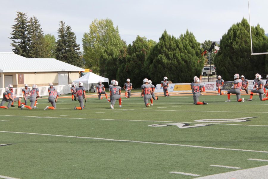 Players in gray and orange warm up and stretch on a green football field.
