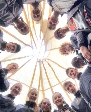 Athletes circle inside a Teepee wearing matching warm-up uniforms. They stare into the camera leaving a glowing orb of light above their heads from the sun entering said teepee above them.