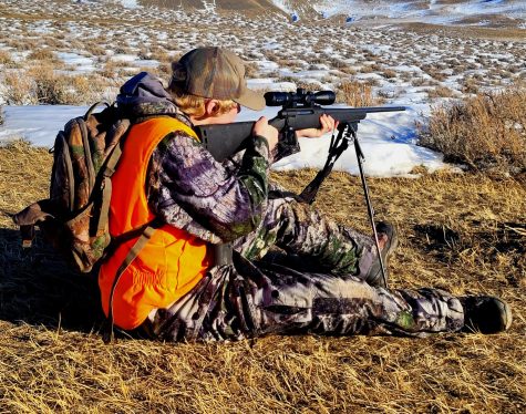 The author looks through the scope on a rifle, while dressed in camo, near a sagebrush hill. He is wearing a fluorescent orange vest to indicate to others that he is there.